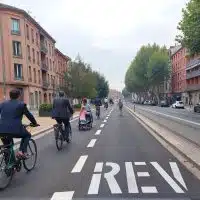 Cyclistes circulant sur une piste du réseau express vélo à Toulouse, reconnaissable à son marquage REV au sol, au cœur d’une avenue urbaine bordée d’immeubles en briques.